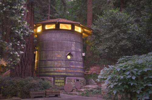 A unique round wooden structure surrounded by tall trees and lush greenery, illuminated softly at dusk.