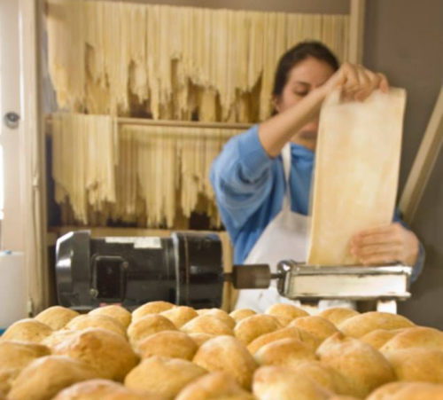 A baker rolls out dough in a bakery, with freshly baked bread rolls in the foreground.