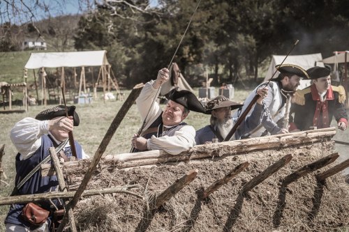 Historical reenactors in period costumes work together to build a wooden structure outdoors, surrounded by tents.