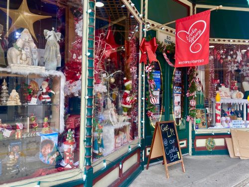 A festive storefront decorated with holiday items, including ornaments, wreaths, and a red banner inviting customers inside.