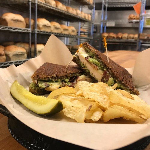 A sandwich with layers of ingredients, served with potato chips and a pickle slice, on a paper-lined basket.