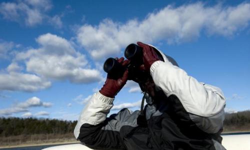 A person in a jacket and gloves uses binoculars against a backdrop of blue sky and clouds.