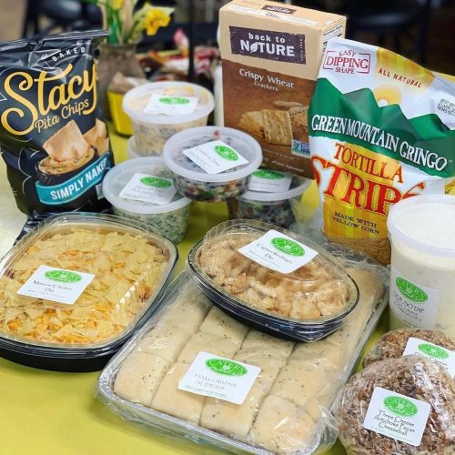 A variety of snacks including pita chips, crispy treats, and dips arranged on a table with flowers in the background.