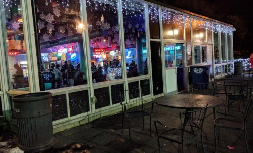 A brightly lit bar with festive decorations, visible through large windows, surrounded by empty outdoor tables.