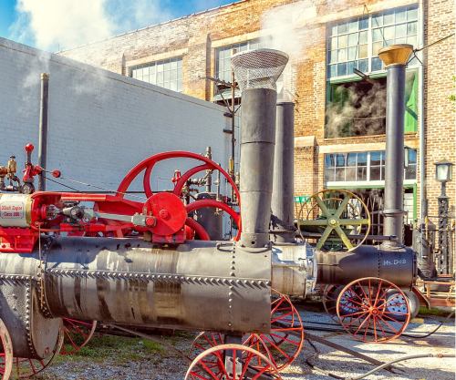 Historic steam engines with red wheels and smokestacks, set against a brick building with large windows.