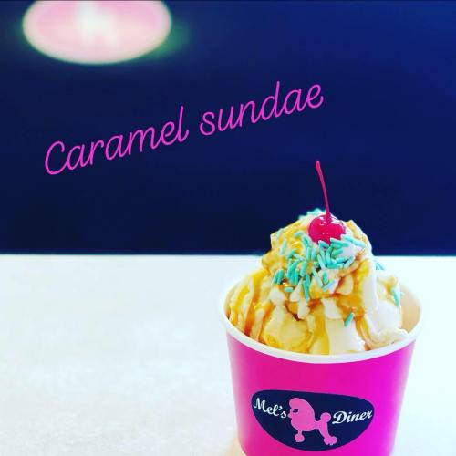 A caramel sundae topped with sprinkles in a pink cup from Mel's Diner, set against a soft-focus background.