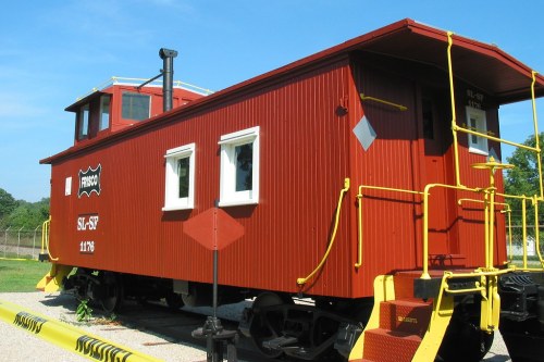 A red caboose with white windows and yellow railings, parked on a gravel area under a clear blue sky.