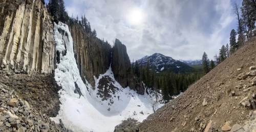 A panoramic view of a snowy landscape featuring a frozen waterfall, rocky cliffs, and distant mountains under a cloudy sky.