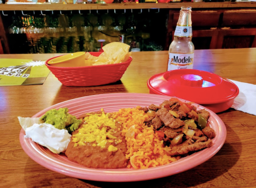 A colorful plate of Mexican food with rice, beans, guacamole, sour cream, and grilled meat, alongside a beer and chips.