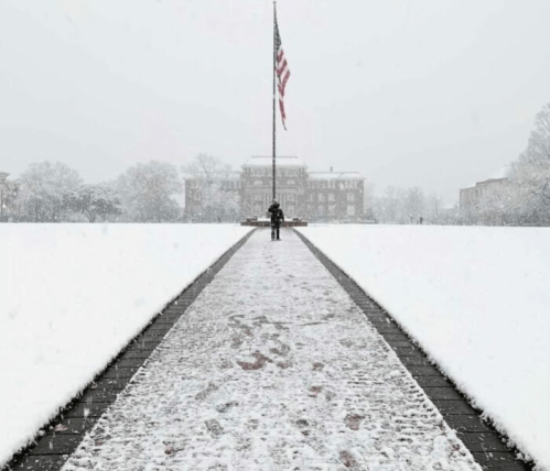 A person stands on a snowy path in front of a flagpole, with a building visible in the background during a snowstorm.