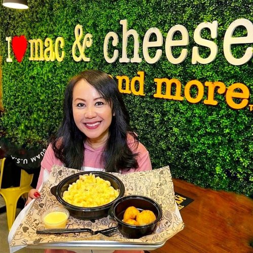 A smiling woman holds a tray with mac and cheese and chicken bites in front of a green wall with "I love mac & cheese" text.