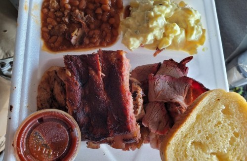 A takeout plate featuring barbecue meats, baked beans, potato salad, and a slice of bread, with a side of sauce.