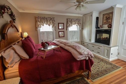 Cozy bedroom with a wooden bed, red bedding, a fireplace, and large windows adorned with floral curtains.