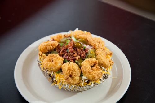 A loaded baked potato topped with fried shrimp, bacon, cheese, jalapeños, and green onions on a white plate.