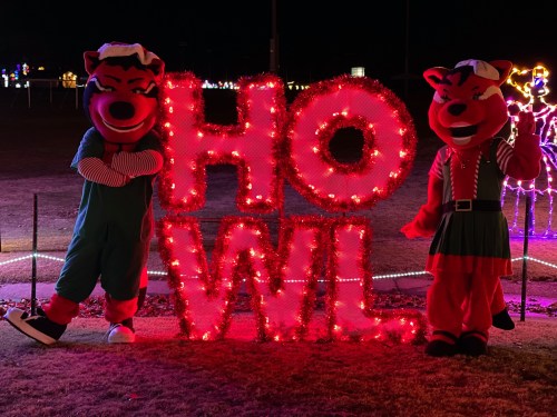 Two costumed characters stand beside a large, illuminated sign that reads "HOWL" in festive lights at night.