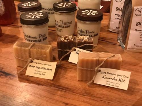 A variety of natural soaps and candles displayed on a wooden table, labeled with their scents and ingredients.