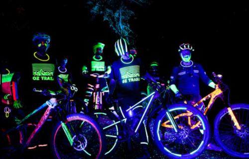 A group of mountain bikers in neon gear and glowing accessories pose together in a dark setting.