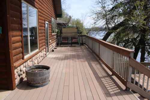 A wooden deck beside a log cabin, overlooking a lake with trees in the background. A planter sits on the deck.