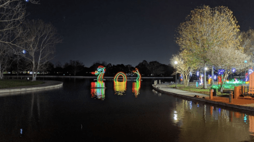 Colorful illuminated decorations reflect on a calm lake at night, surrounded by trees and pathways.