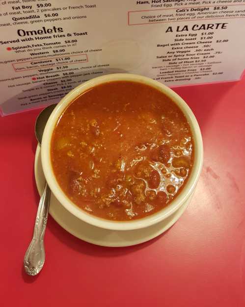 A bowl of hearty tomato soup with chunks of meat, served on a pink table next to a menu.