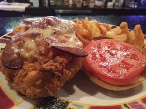 A plate featuring a fried chicken sandwich with melted cheese, crispy bacon, fresh tomato slices, and a side of fries.