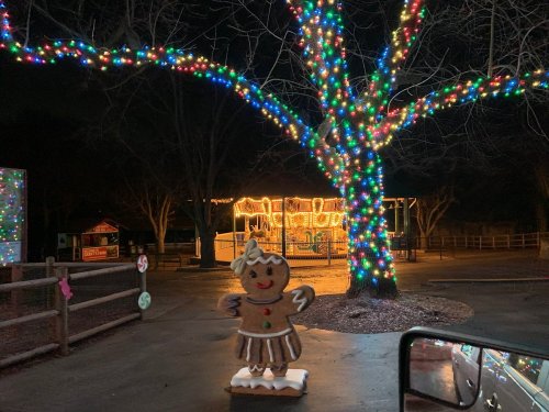 A gingerbread figure stands in front of a brightly lit tree and carousel at night, surrounded by festive decorations.