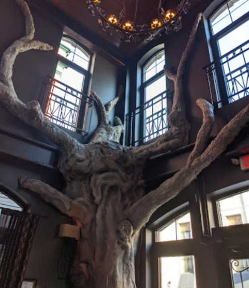 A large, artistic tree sculpture with twisted branches inside a room with large windows and a chandelier.