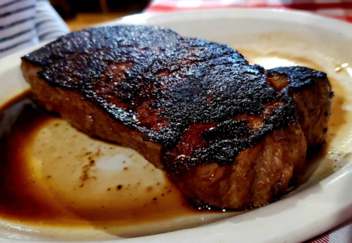 A well-cooked steak with a charred exterior, resting in a shallow pool of sauce on a white plate.