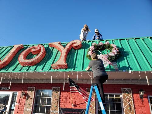 Two people decorate a building rooftop with large letters spelling "JOY" while another person adjusts a wreath on a ladder.