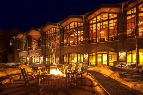 A cozy outdoor fire pit surrounded by wooden chairs, with a warmly lit lodge in the background at night.