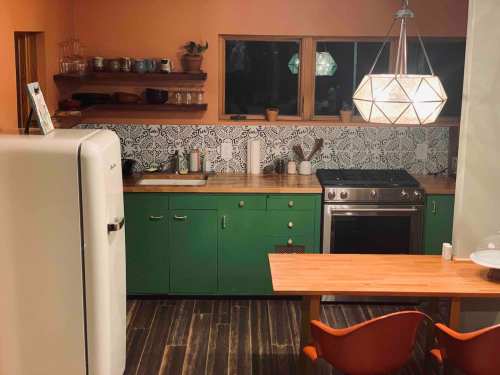 A modern kitchen featuring green cabinets, a white fridge, a wooden table, and a geometric pendant light.