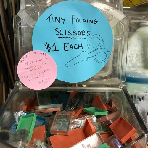 A container of tiny folding scissors for sale at $1 each, with a note advising against testing utensils.