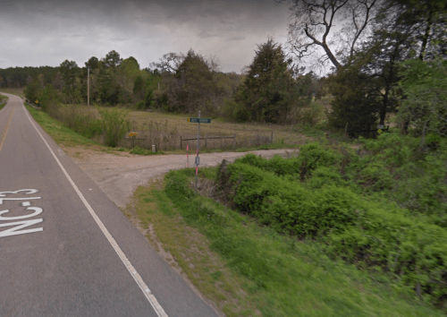 A rural road intersection with a gravel path, surrounded by greenery and trees, under a cloudy sky.