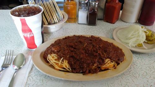 A plate of spaghetti topped with chili, served with a drink and sides of onions and pickles on a retro diner table.