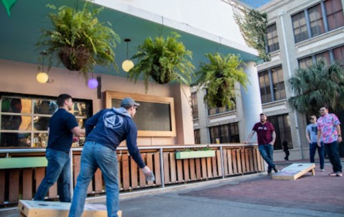 A group of people playing cornhole outside a bar with hanging plants and a sunny atmosphere.