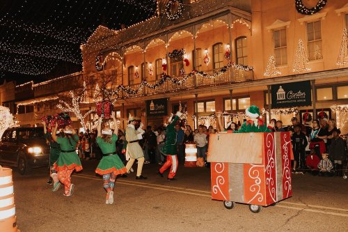 A festive parade scene with performers in colorful costumes dancing in front of a beautifully lit building.