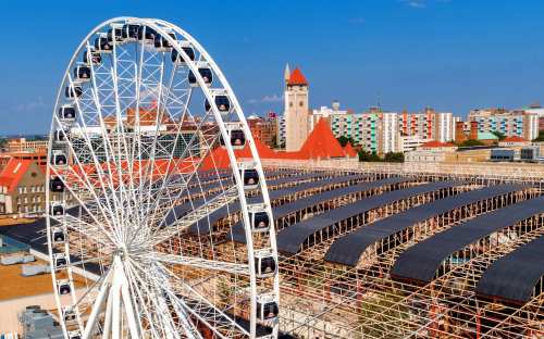 Aerial view of a large Ferris wheel near a bustling cityscape with colorful buildings and a clock tower in the background.