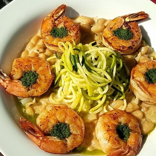 A plate of shrimp on a bed of white beans, topped with green sauce and served with spiralized zucchini noodles.