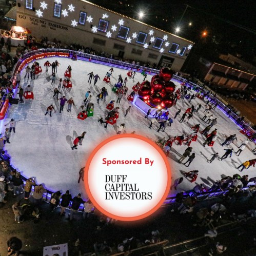 Aerial view of a festive ice skating rink with people skating, surrounded by holiday decorations and lights.