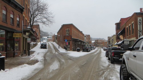 Snow-covered street in a historic town, featuring brick buildings, shops, and a steep hill in the background.