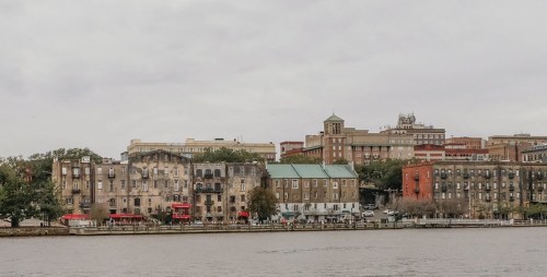 A waterfront view of historic buildings and restaurants along a river, under a cloudy sky.