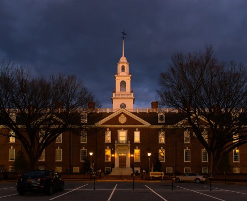 Historic building illuminated at dusk, featuring a clock tower and surrounded by bare trees and parked cars.