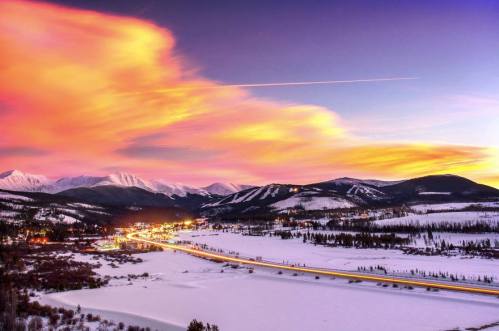 A stunning winter landscape at sunset, featuring snow-covered mountains, a river, and colorful clouds in the sky.