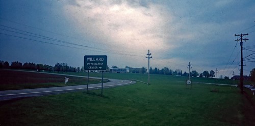 Sign for Willard Psychiatric Center along a winding road, with a cloudy sky and grassy fields in the background.
