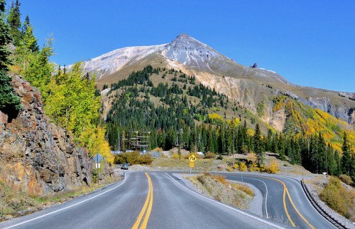 A winding road leads through vibrant autumn trees, with a snow-capped mountain in the background under a clear blue sky.