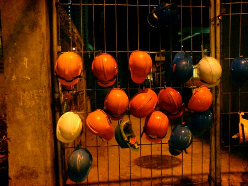 A fence with various colored hard hats hanging on it, illuminated by dim light at night.