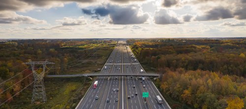 Aerial view of a busy highway surrounded by autumn foliage and cloudy skies. Power lines run alongside the road.