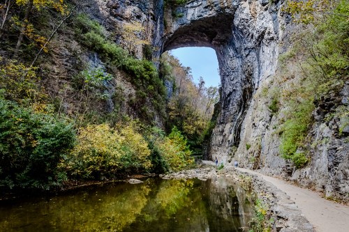 A natural stone arch towers over a serene river, surrounded by lush greenery and rocky cliffs in a tranquil landscape.