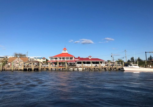 A waterfront restaurant with a red roof and wooden docks, surrounded by blue skies and calm water.