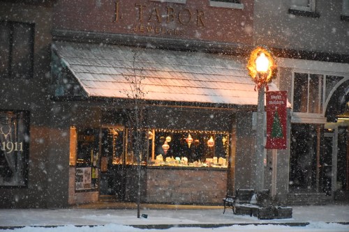 A jewelry store window decorated with lights, surrounded by falling snow and a festive street lamp.